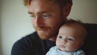 Tender moment between young father with ginger beard holding baby in casual home setting, soft natural window lighting creates intimate atmosphere for family portrait.