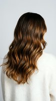 Woman with long wavy brown hair with caramel highlights wearing white sweater from behind showing healthy hair texture and color for beauty salon.