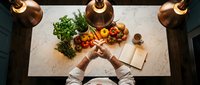 Chef in white uniform planning a recipe at a marble kitchen counter with fresh vegetables, herbs, and an open cookbook under warm copper pendant lights.