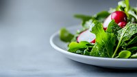 Fresh green salad with radish and arugula leaves on white ceramic plate against gray background, selective focus with copy space for menu design.