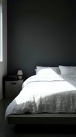 Minimalist bedroom interior with white bedding and gray pillows against dark charcoal wall, featuring small wooden nightstand with lamp and natural light from window for home decor and interior design projects.