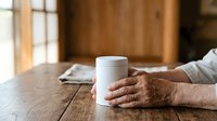 Senior man with weathered hands holding white ceramic mug on wooden table indoors. Suitable for lifestyle, wellness, and beverage marketing campaigns.