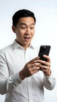 Asian male professional in white button-up shirt holding smartphone, displaying excited expression while viewing screen content against white background for business and technology concepts.