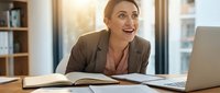 Happy blonde businesswoman smiling while working at desk with laptop and books in bright modern office environment for professional success concepts.