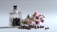Small glass bottle with black peppercorns and cork stopper, scattered peppercorns on white surface, pink flowers in background.