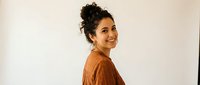 Young Hispanic woman with curly hair in messy bun wearing brown sweater smiling over shoulder against neutral background for lifestyle content.