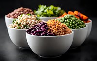 Assorted legumes and vegetables in white ceramic bowls on dark background. Fresh ingredients for healthy vegetarian cooking and protein-rich diet.