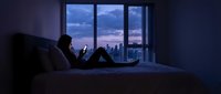 Young woman relaxing on bed using smartphone at twilight with city skyline view through large windows in modern apartment bedroom interior.