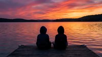 Two children sitting on wooden dock watching dramatic sunset over calm lake with vibrant orange and pink sky reflecting on water surface creating peaceful moment.