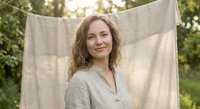 Young Caucasian woman with curly hair smiling outdoors near hanging linen fabric in natural sunlight for lifestyle and wellness concepts.