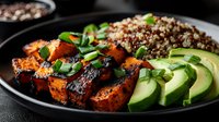 Grilled spicy chicken pieces served with quinoa, fresh sliced avocado and green onions on black plate, healthy protein-rich meal against dark background.