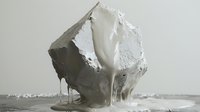 White liquid paint pouring over rough geometric stone or concrete shape creating drips and flows against neutral background, monochromatic minimal art composition.