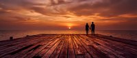 Silhouetted couple standing on wooden pier at sunset, holding hands while watching golden sun descend over calm ocean horizon during romantic evening. Perfect for travel and relationship themes.