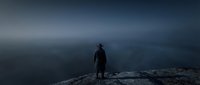 Man in dark coat standing alone on rocky mountain peak overlooking misty valley landscape at dusk. Solitude and contemplation concept for editorial and lifestyle use.