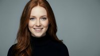 Smiling redhead woman in black turtleneck sweater against gray background, professional headshot portrait for business and corporate marketing materials.