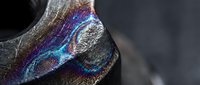 Iridescent metallic surface with rainbow holographic reflections showing purple blue and gold colors on dark textured material with dramatic lighting effects.