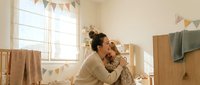 Young mother with dark hair in bun embracing toddler daughter in cozy nursery room with wooden furniture, bunting flags and soft natural lighting.