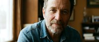 Middle-aged Caucasian man with gray beard wearing denim shirt, serious expression, indoor setting with warm lighting, suitable for business, lifestyle, and portrait projects.