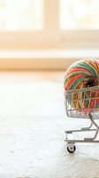 Colorful yarn ball in miniature shopping cart on neutral surface with blurred interior background, representing craft supplies, hobbies, and retail shopping concepts.