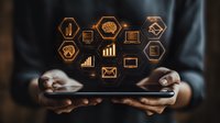 Digital business icons floating over tablet in hands showing analytics, communication and productivity tools. Orange glowing symbols on dark background.