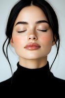 Young Asian woman with closed eyes wearing black turtleneck, natural makeup, freckles and glossy lips against light background. Clean beauty portrait for cosmetic advertising.