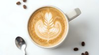 Overhead view of cappuccino with latte art foam design in white ceramic mug, surrounded by coffee beans and spoon on white surface. Perfect for café, coffee shop, beverage, and hospitality marketing.