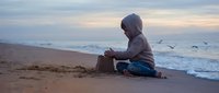 Young child in hoodie building sandcastle on beach at sunset with seagulls flying overhead and gentle waves washing shore creating peaceful coastal scene.
