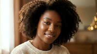 Young African American woman with natural curly hair smiling warmly in cozy beige sweater. Professional portrait with soft lighting for lifestyle content.