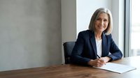 Mature woman with gray hair, Caucasian ethnicity, wearing dark blazer and white shirt, sitting at wooden desk writing on document in modern office with concrete wall and window.