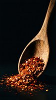 Wooden spoon with crushed red pepper flakes spilling onto dark surface creating dramatic lighting effect for culinary and cooking concepts.