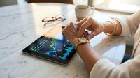 Professional businesswoman analyzing financial data and stock market charts on digital tablet at modern office desk with coffee and eyeglasses nearby.