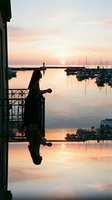 Woman silhouette sitting on balcony railing overlooking serene harbor at sunset with moored sailboats, calm water reflections, and golden sky for travel, lifestyle, and contemplation themes.