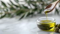 Wooden spoon pouring fresh extra virgin olive oil into glass bowl with green olives and leaves on white marble background, natural light and copy space.