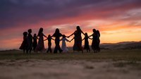 Group of diverse people holding hands in circle dancing together at sunset with dramatic colorful sky and mountain landscape background for teamwork concepts.