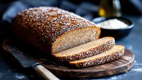 Freshly baked whole grain bread loaf with sesame seeds on wooden cutting board, sliced to show soft texture inside. Dark moody food photography.