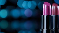 Vibrant pink lipstick against blurred blue bokeh background, closeup macro shot showing metallic texture and shimmer finish in cosmetic product photography.