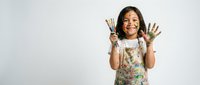 Young Asian girl artist holding paintbrushes with paint-covered hands showing creative mess from art class activities on white background.