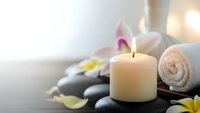Spa wellness scene with burning candle, rolled towel, smooth stones and tropical flowers creating peaceful relaxation atmosphere for massage therapy.