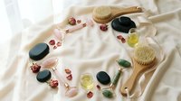 Spa wellness flat lay with hot stones, essential oils, jade roller, dry brush and rose petals on white fabric background for relaxation and self-care routine.