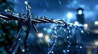 Water drops falling on barbed wire fence during night rain storm, creating dramatic splash effect with bokeh lights and deep blue atmospheric mood.