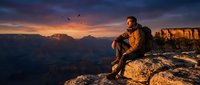 Man in casual outdoor clothing sitting on rocky cliff edge overlooking vast canyon landscape at golden hour sunset, contemplating nature scenery for travel and adventure concepts.
