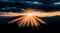 Dramatic sunburst breaks through dark storm clouds over mountain silhouettes at sunset, creating golden rays of light stretching across moody evening sky.
