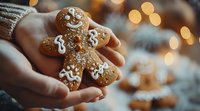 Hands holding freshly baked gingerbread man cookie decorated with white icing and sugar sprinkles against warm bokeh lights background, festive holiday mood.