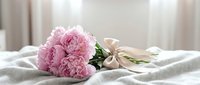 Pink peony bouquet tied with ribbon on white linen bedding in soft natural light for romantic wedding decor and floral arrangements.