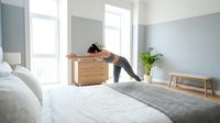 Woman stretching over wooden dresser in bright bedroom with natural light, wearing casual athletic wear. Wellness and morning routine concept for home decor and lifestyle content.