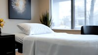 Clean white massage table with fresh linens and pillow in modern spa treatment room featuring natural light and decorative sunflowers, ready for wellness session.