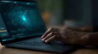 Dark workspace with hands typing on laptop keyboard displaying glowing blue digital code and data visualization on screen, moody tech atmosphere.
