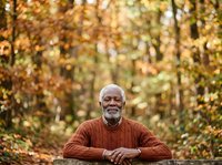 Senior African American man with gray beard wearing rust-colored sweater, seated outdoors among autumn foliage. Suitable for lifestyle, wellness, and seasonal editorial use.