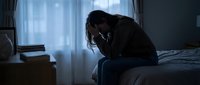 Depressed young woman sitting on bed edge in dark bedroom holding head in hands showing mental health struggles and emotional pain.