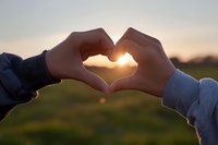 Hands forming heart shape against sunset sky over green field landscape capturing love romance and connection outdoors during golden hour.